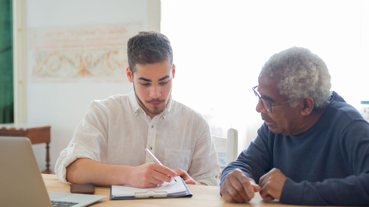 An older man at a sunlit table beside a younger visitor who is filling in paperwork on a clipboard.