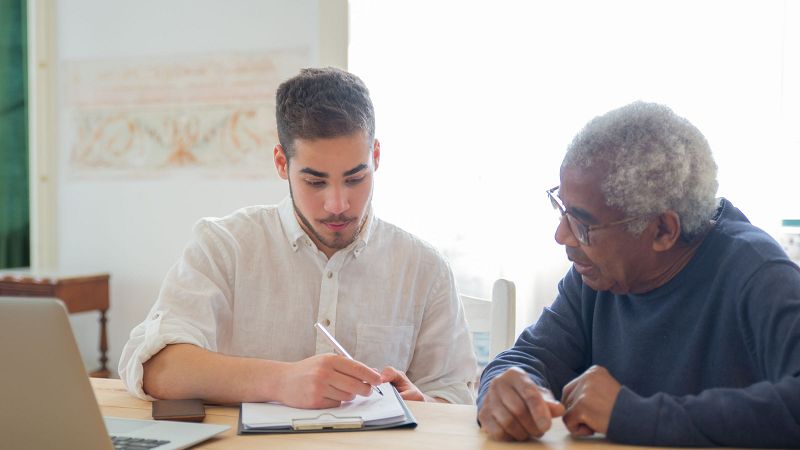 An older man at a sunlit table beside a younger visitor who is filling in paperwork on a clipboard.