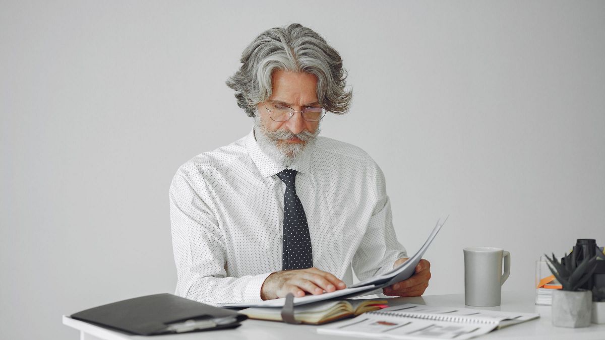 Un hombre con lentes revisa con calma una carpeta de páginas impresas en un escritorio blanco.