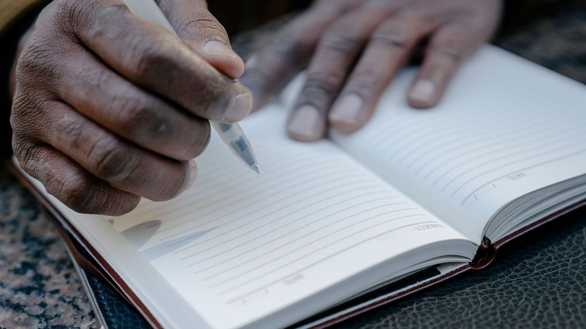 A hand writing an entry into an open bound ledger with ruled lines, resting on a leather folio.