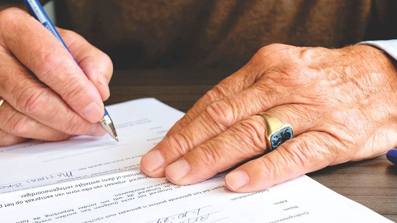 An older person signing a document, with someone assisting at the table.